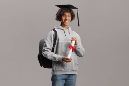 Foto de Joven estudiante afroamericano masculino con una mochila que sostiene un título universitario aislado sobre fondo gris - Imagen libre de derechos