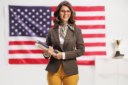 Photo for Young female teacher with glasses holding books with USA flag in the background - Royalty Free Image