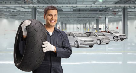 Photo for Auto mechanic worker holding a tire inside a garage with cars - Royalty Free Image