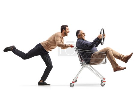 Photo for Guy pushing a mature man inside a shopping cart holding a steering wheel isolated on white background - Royalty Free Image