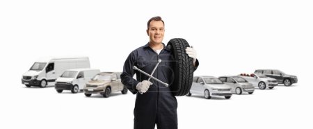 Téléchargez les photos : Mécanicien automobile avec de nombreux véhicules portant un pneu et tenant un outil de clé isolé sur fond blanc - en image libre de droit