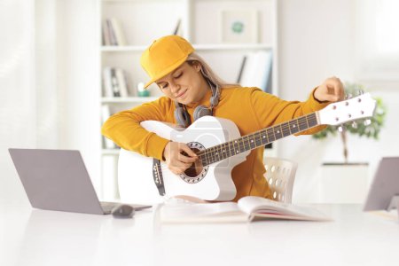 Téléchargez les photos : Jeune femme accordant une guitare acoustique devant un ordinateur portable à la maison - en image libre de droit