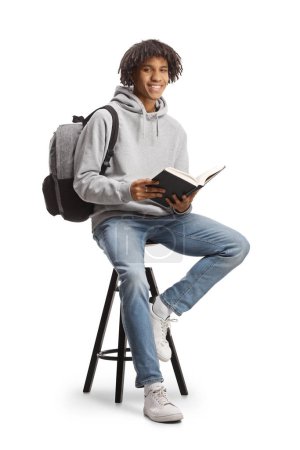 Foto de Estudiante afroamericano con una mochila sentada en una silla y sosteniendo un libro abierto aislado sobre fondo blanco - Imagen libre de derechos