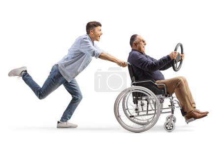 Photo for Full length profile shot of a guy pushing a mature man in a wheelchair holding a steering wheel isolated on white background - Royalty Free Image
