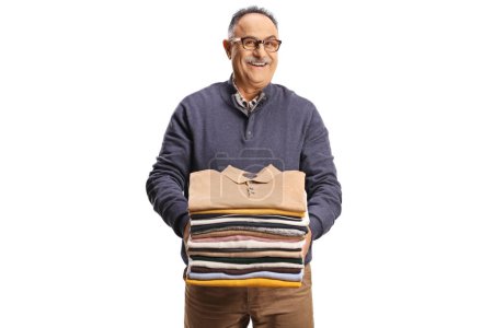 Foto de Alegre hombre maduro sosteniendo una pila de ropa doblada y sonriendo a la cámara aislada sobre un fondo blanco - Imagen libre de derechos