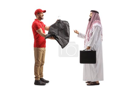 Téléchargez les photos : Travailleur livrant des vêtements de nettoyage à sec à un homme arabe en vêtements ethniques isolé sur un dosseret blanc - en image libre de droit