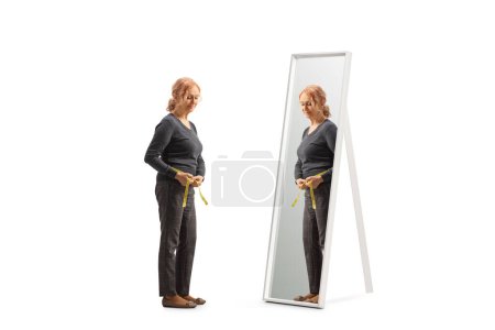 Photo for Full length shot of a pensive mature woman measuring waist in front of a mirror isolated on white background - Royalty Free Image