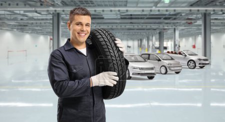 Téléchargez les photos : Travailleur mécanicien automobile portant un pneu à l'épaule et souriant à la caméra à l'intérieur d'un garage - en image libre de droit