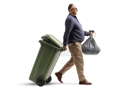 Foto de Foto de perfil completo de un hombre maduro caminando con una bolsa de residuos y tirando de un contenedor de plástico aislado sobre fondo blanco - Imagen libre de derechos