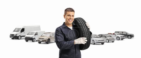Téléchargez les photos : Travailleur mécanicien automobile avec de nombreux véhicules portant un pneu à l'épaule et souriant à la caméra isolée sur fond blanc - en image libre de droit