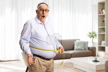 Photo for Shocked mature man measuring waist with a tape in a living room at home - Royalty Free Image