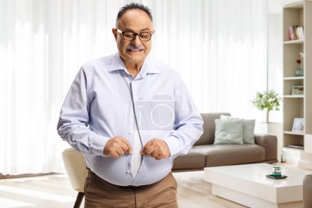 Photo for Mature man with a big belly trying to button a tight shirt at home - Royalty Free Image