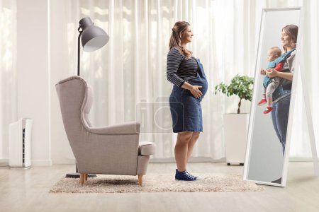 Photo for Full length shot of a pregnant woman looking herself at a mirror and imagining her baby at home - Royalty Free Image