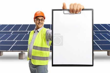 Téléchargez les photos : Homme ingénieur mature dans un gilet réfléchissant montrant un document vierge sur un presse-papiers devant des panneaux solaires isolés sur fond blanc - en image libre de droit