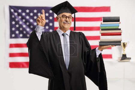 Photo for Smiling mature man in a graduation gown holding a pile of books and pointing in front of a USA flag - Royalty Free Image