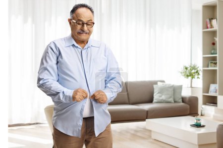Téléchargez les photos : Mature homme essayant de boutonner une petite chemise dans un salon à la maison - en image libre de droit