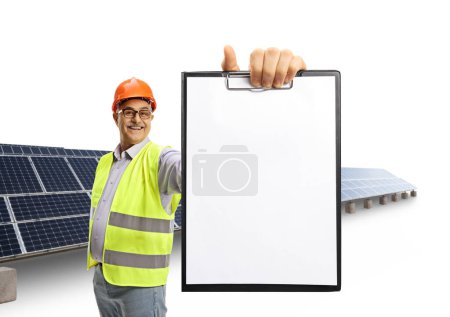 Téléchargez les photos : Joyeux ingénieur mâle mature dans un gilet réfléchissant tenant un presse-papiers devant le champ solaire isolé sur fond blanc - en image libre de droit