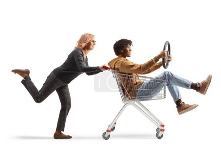 Photo for Woman pushing a young african american man inside a shopping cart sitting and holding a steering wheel isolated on white background - Royalty Free Image