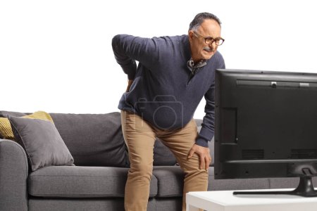 Téléchargez les photos : Homme mûr avec mal de dos se lever d'un canapé devant la télévision isolé sur fond blanc - en image libre de droit
