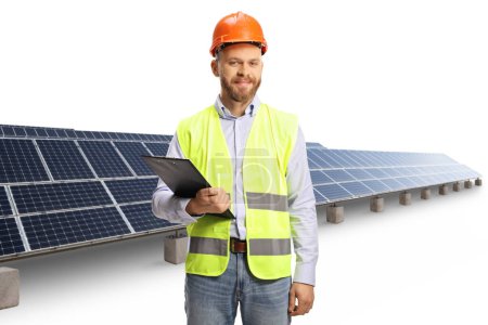 Téléchargez les photos : Ingénieur avec un casque de sécurité tenant un presse-papiers devant la centrale photovoltaïque isolé sur fond blanc - en image libre de droit