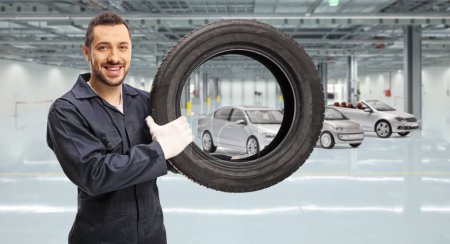 Téléchargez les photos : Mécanicien automobile tenant un pneu de voiture et souriant à la caméra à l'intérieur d'un garage - en image libre de droit