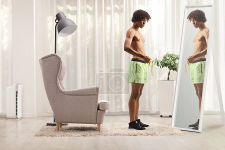 Photo for African american male athlete measuring waist in front of a mirror in a roo - Royalty Free Image