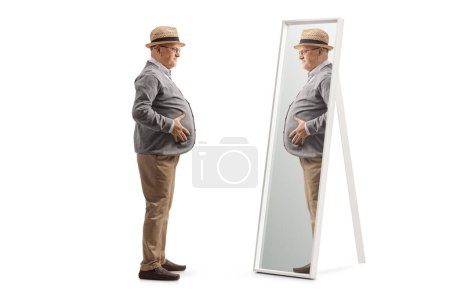 Téléchargez les photos : Plan de profil complet d'un homme âgé avec un gros ventre debout devant un miroir isolé sur fond blanc - en image libre de droit
