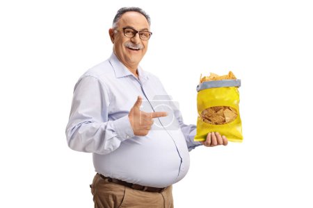 Foto de Alegre hombre maduro sosteniendo una bolsa de patatas fritas de tortilla y apuntando aislado sobre fondo blanco - Imagen libre de derechos