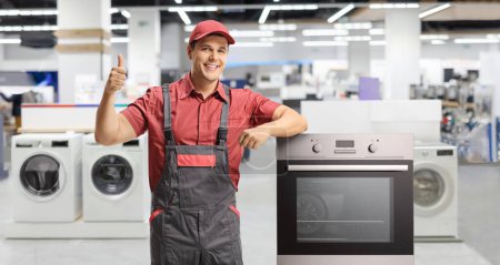 Foto de Reparador en uniforme de pie junto a un horno eléctrico y dando pulgares hacia arriba dentro de una tienda - Imagen libre de derechos