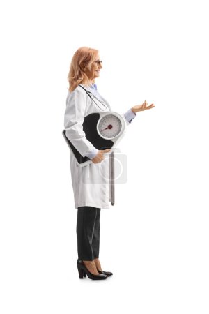 Téléchargez les photos : Prise de vue complète du profil d'une femme médecin tenant une balance de poids et faisant un geste avec la main isolée sur fond blanc - en image libre de droit