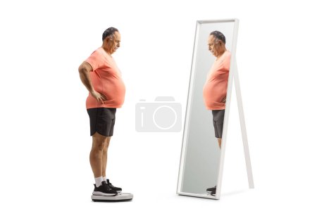 Téléchargez les photos : Homme mûr avec un gros ventre debout sur une échelle de poids et regardant un miroir isolé sur fond blanc - en image libre de droit