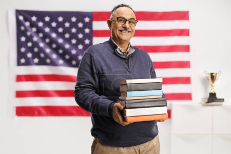 Photo for Smiling mature man holding a pile of books and posing in front of the USA flag hanging on wall - Royalty Free Image