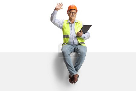Foto de Retrato de cuerpo entero de un ingeniero varón maduro sentado en un panel en blanco y saludando a la cámara aislado sobre fondo blanco - Imagen libre de derechos