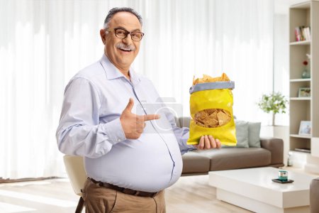 Photo for Cheerful mature man holding a pack of tortilla chips and pointing at home in a living room - Royalty Free Image