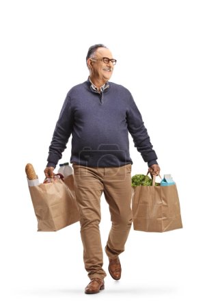 Photo for Full length portrait of a mature man carrying grocery bags and walking towards camera with his head to the side isolated on white background - Royalty Free Image