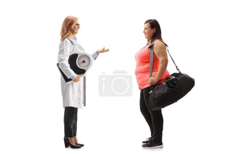 Foto de Foto de perfil completo de una doctora que sostiene una báscula de peso y habla con una mujer joven con sobrepeso aislada sobre fondo blanco - Imagen libre de derechos