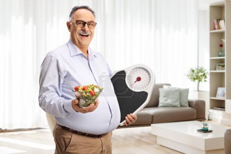 Photo for Mature man holding a salad and a weight scale at home in a living room - Royalty Free Image