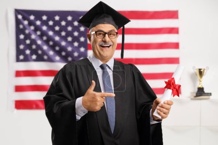 Photo for Mature man in a graduation gown holding a diploma and pointing in front of USA flag - Royalty Free Image