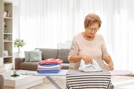 Photo for Elderly woman ironing clothes at home in a living room - Royalty Free Image