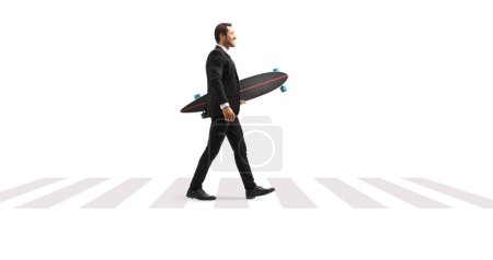 Photo for Full length profile shot of a businessman in a suit walking at a pedestrian crossing and holding a longboard isolated on white background - Royalty Free Image
