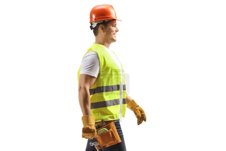 Photo for Profile shot of a construction worker with a helmet and tool belt walking isolated on white background - Royalty Free Image
