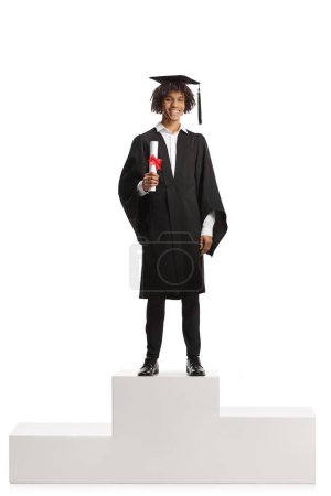 Photo for Male african american student in a graduation gown holding a diploma and standing on a winner pedestal isolated on white background - Royalty Free Image