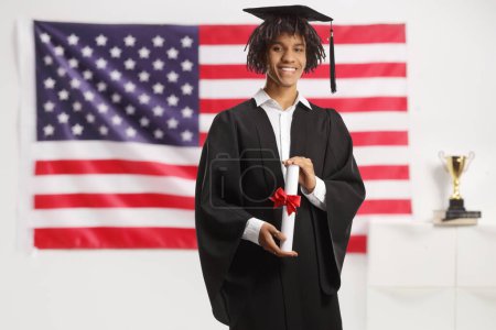 Photo for African american male student in a graduation gown holding a diploma and standing in front of a USA flag - Royalty Free Image