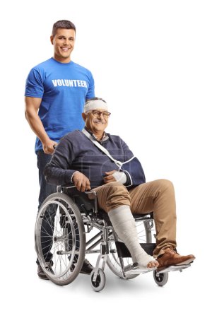 Foto de Voluntario empujando a un hombre maduro con un brazo roto y vendaje en la cabeza en una silla de ruedas aislada sobre fondo blanco - Imagen libre de derechos