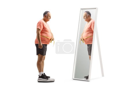 Photo for Mature man measuring waist in front of a mirror and standing on a weight scale isolated on white background - Royalty Free Image