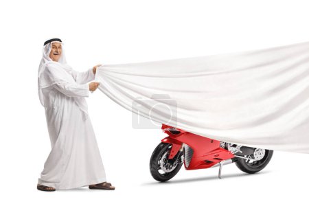 Foto de Hombre árabe con bata tradicional sosteniendo un trozo blanco de tela frente a una moto de carreras roja aislada sobre fondo blanco - Imagen libre de derechos