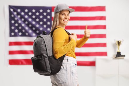 Photo for Female student with a backpack showing thumbs up in front of the USA flag - Royalty Free Image
