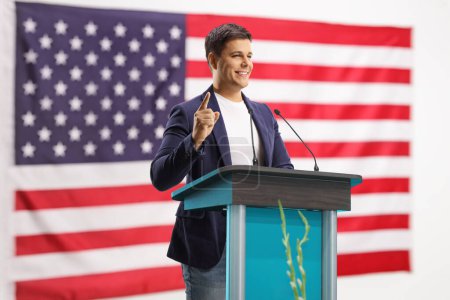 Photo for Young man giving a speech on a pedestal and gesturing with finger with the USA flag behind - Royalty Free Image