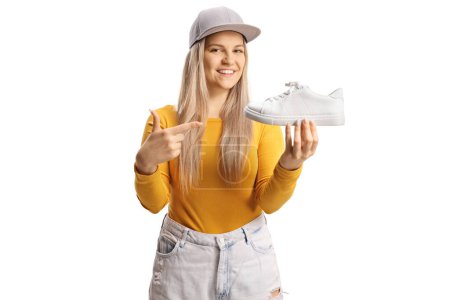 Téléchargez les photos : Tendance casual jeune femme tenant une sneaker blanche et pointant isolé sur fond blanc - en image libre de droit