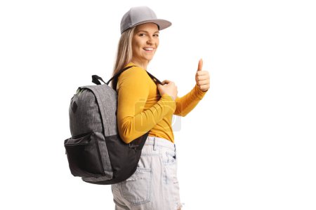 Foto de Estudiante femenina con una mochila mirando por encima del hombro y haciendo gestos con los pulgares hacia arriba aislada sobre fondo blanco - Imagen libre de derechos